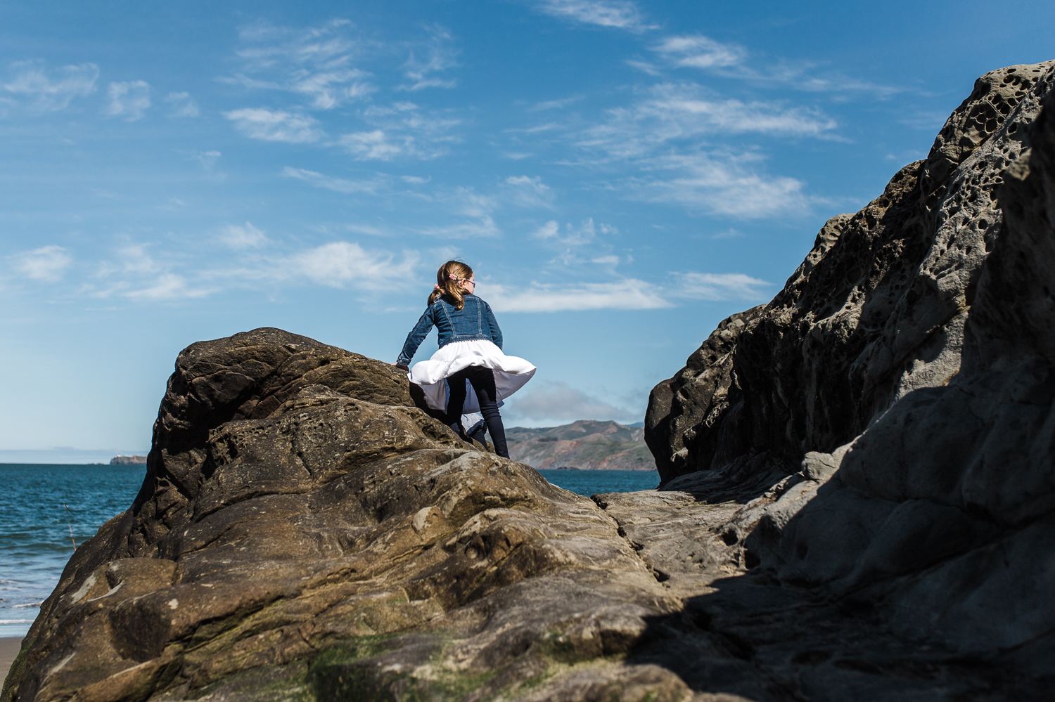 five year old girl climbing on a rock and looking out at the ocean with her dress blowing in the wind at Baker Beach {San Francisco Child Photographer}