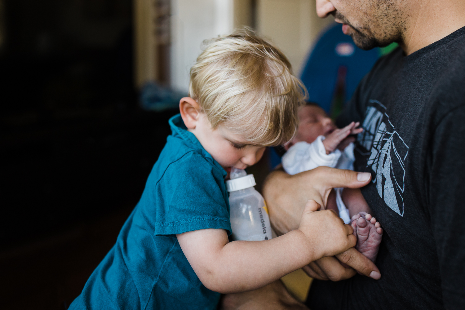 A toddler drinking his bottle and leaning into his dad and looking sad and unsure about his two day old newborn brother in his dad's arms.