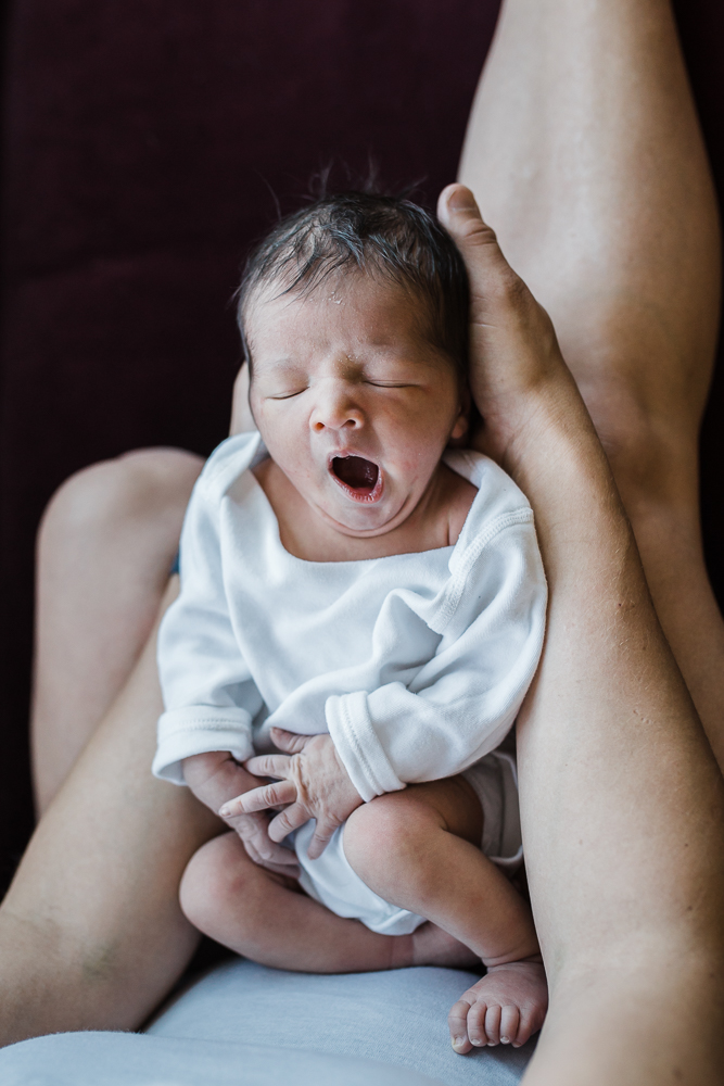 A newborn in a long-sleeved white onesie laying on his mom and yawning with his eyes closed.