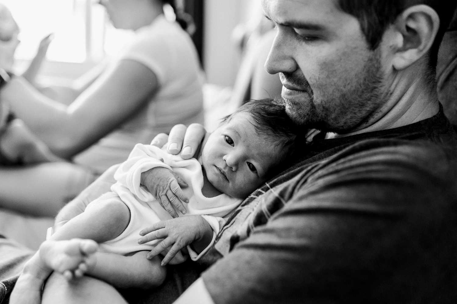 A newborn baby with his eyes open resting on his dad's chest with mom playing with the toddler in the background.