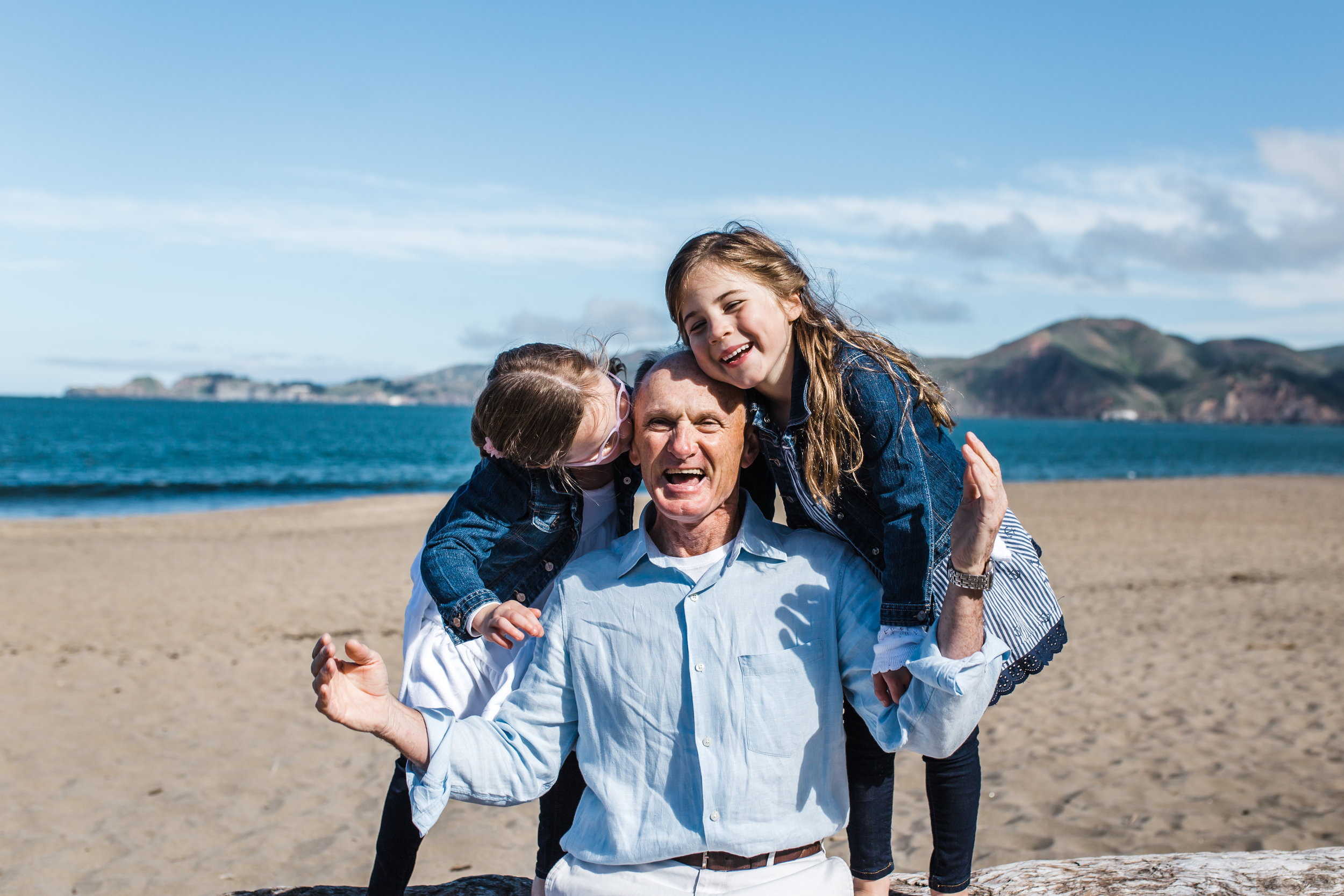 A dad sitting on the beach with his twin five year old girls while the girls each hug and kiss him from either side of him at a Bay Area beach with the pacific ocean in the background.