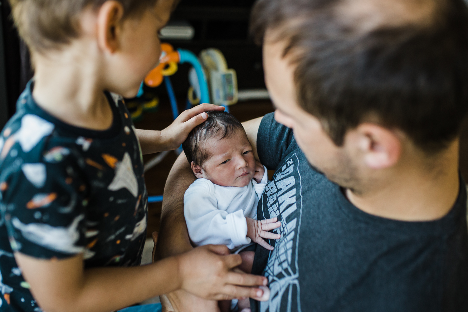 An older brother looking down and gently touching a fresh newborn just two days old who is being held by their dad.