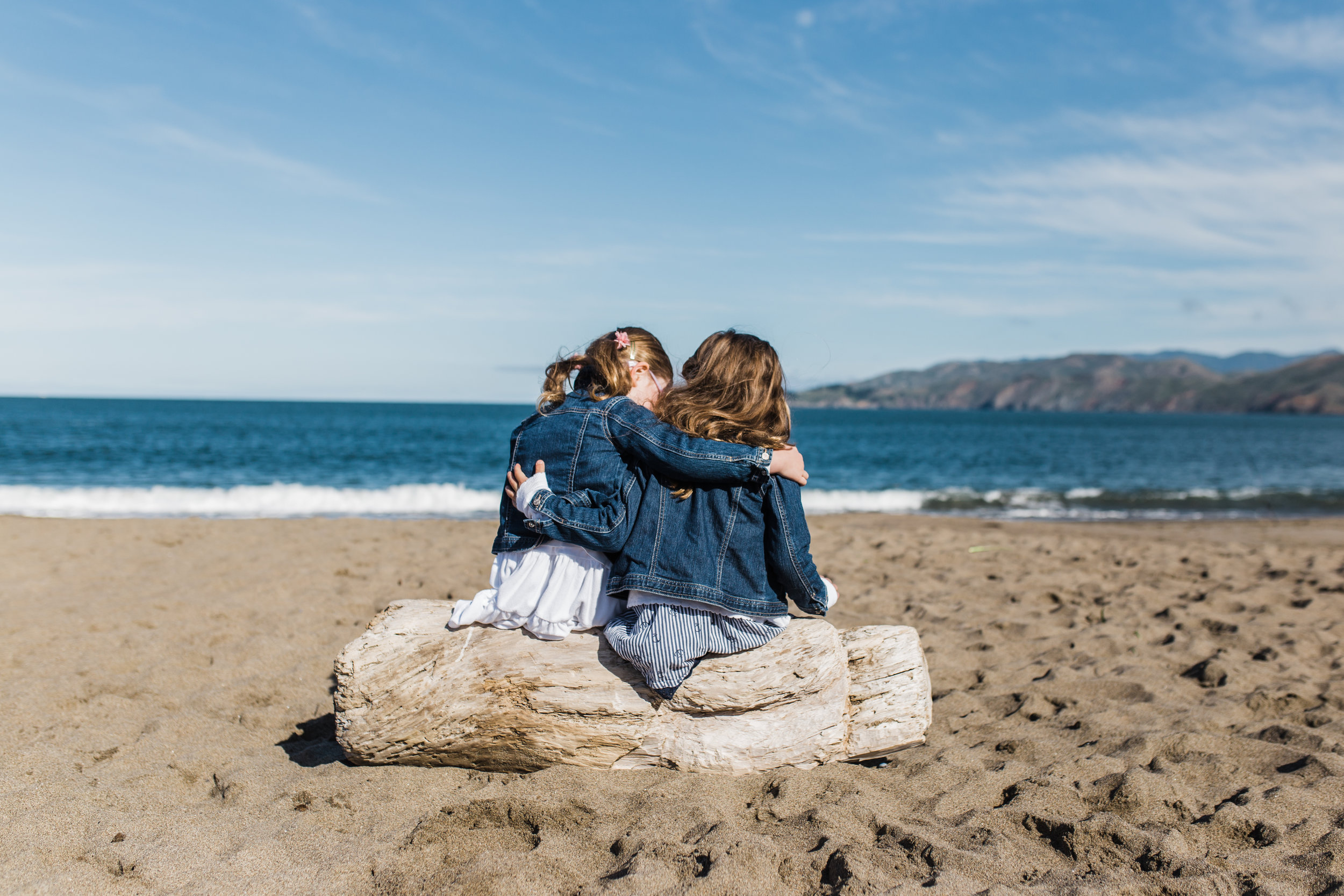 five year old twin sisters sitting on a piece of driftwood on Baker Beach with their arms wrapped around each other 