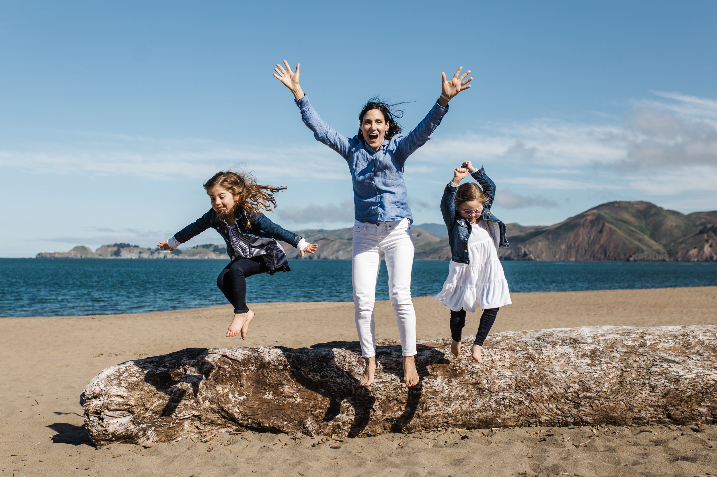 A mom and twin five year old girls jumping off of a large piece of driftwood at Baker beach and smiling