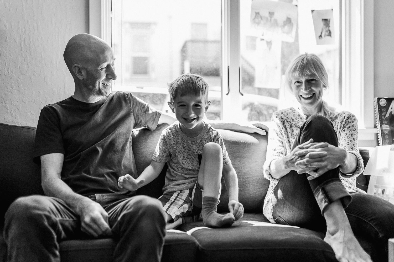 five year old boy sitting with his parents on a couch in front of a window in their house 