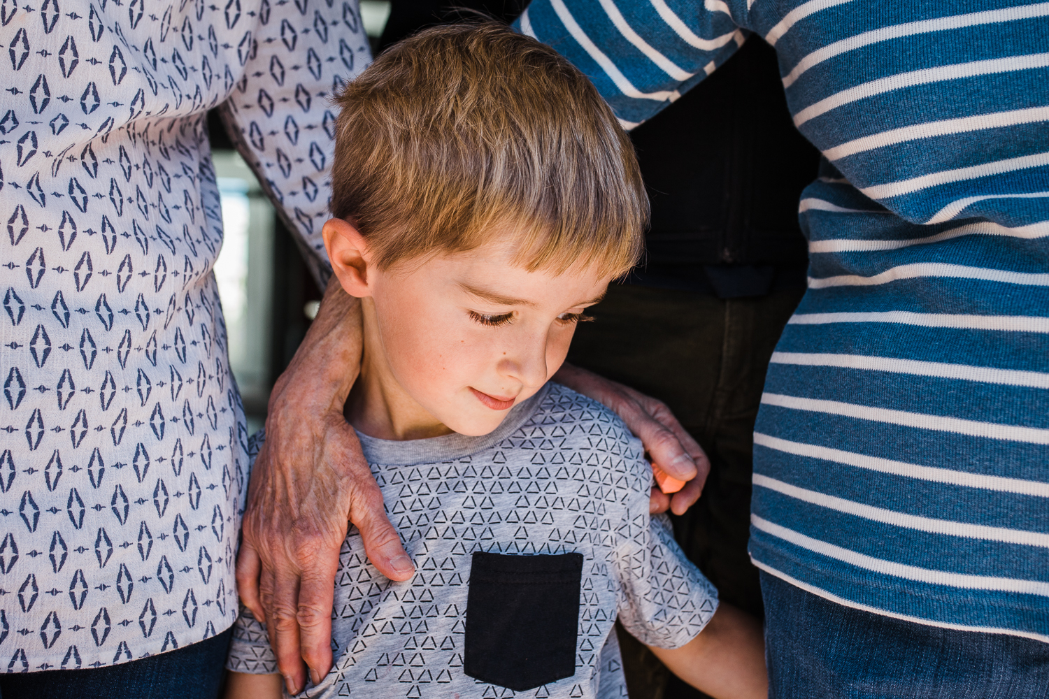 close up of five year old boy standing next to his mother and grandmother with their hands touching his back 