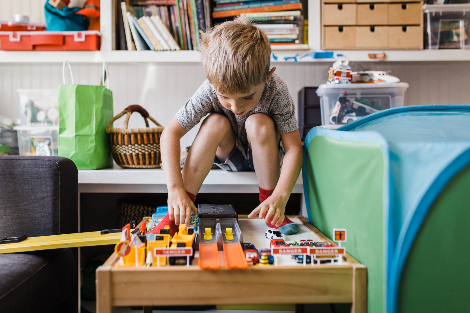 five year old boy sitting on a shelf while playing with his cars - his feet propped up on lego table 