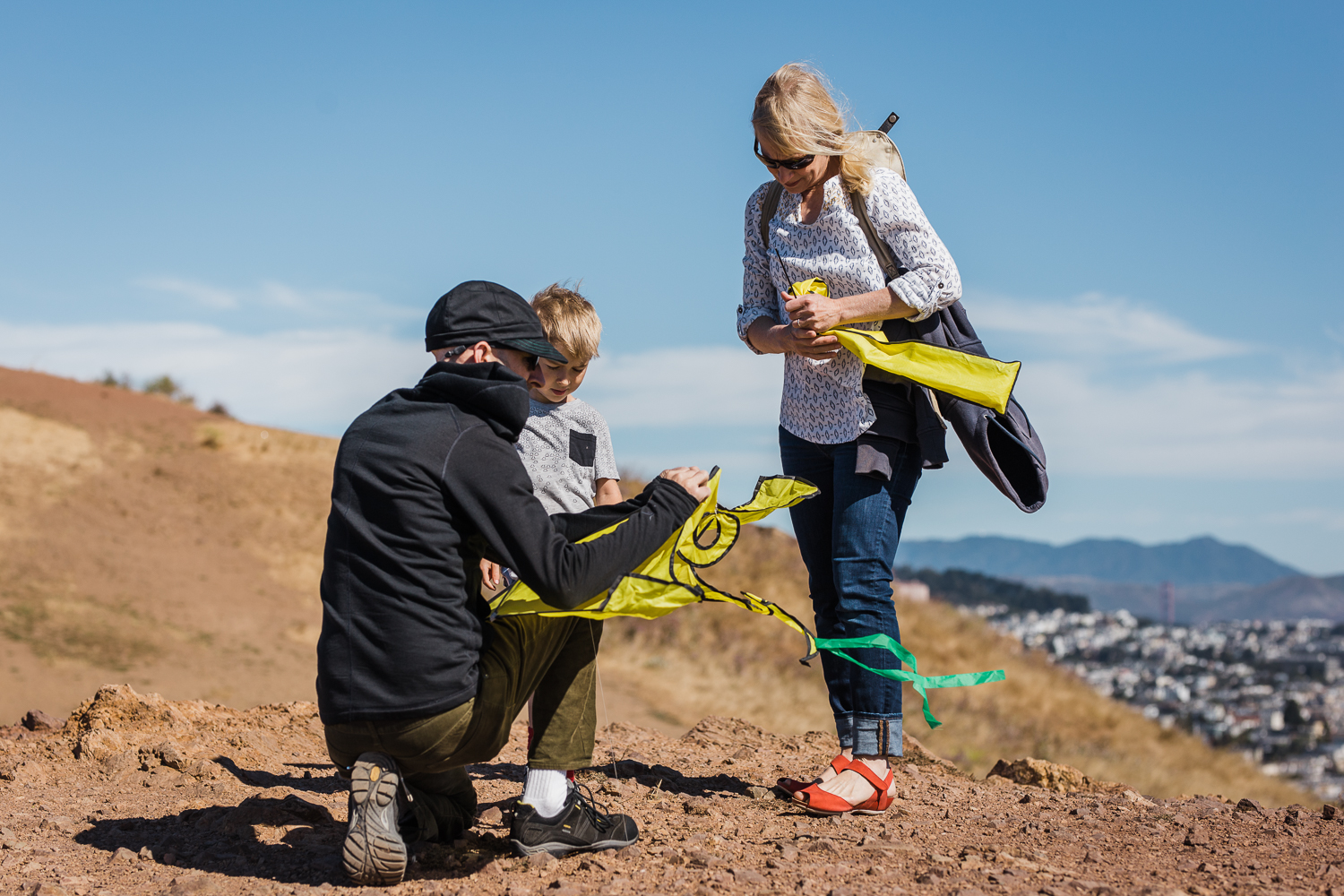 A family of three setting up to fly a kite overlooking the city from Bernal Hill with the Golden Gate Bridge in the distance