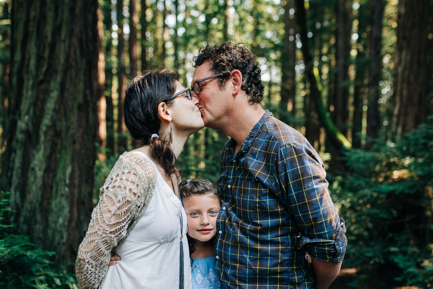 parents kissing as their eight year old daughter squeezes between them while in the woods at Joaquin Miller Park in Oakland