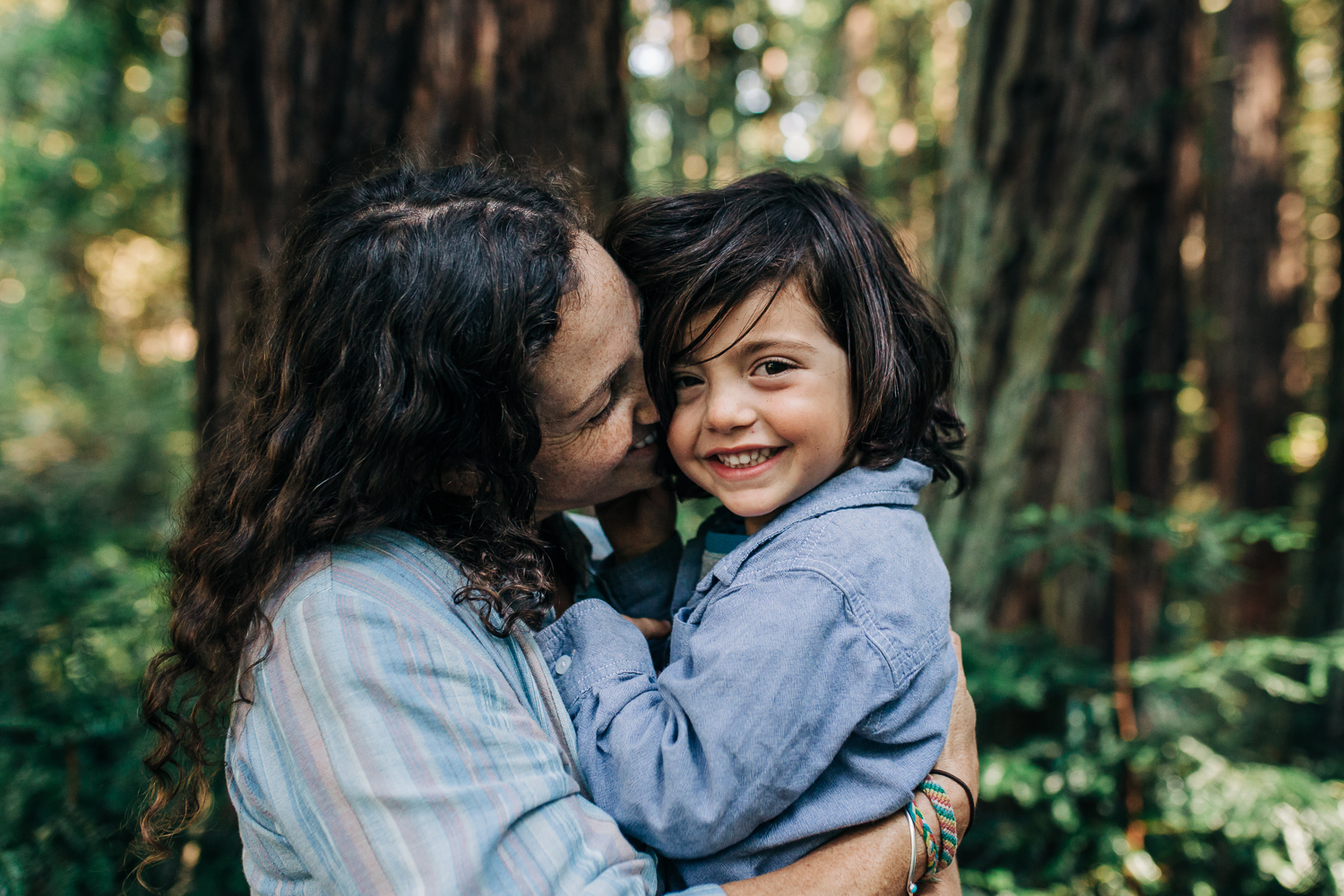 Mom snuggling her four year old boy as he looks at the camera and smiles while in the woods in Oakland