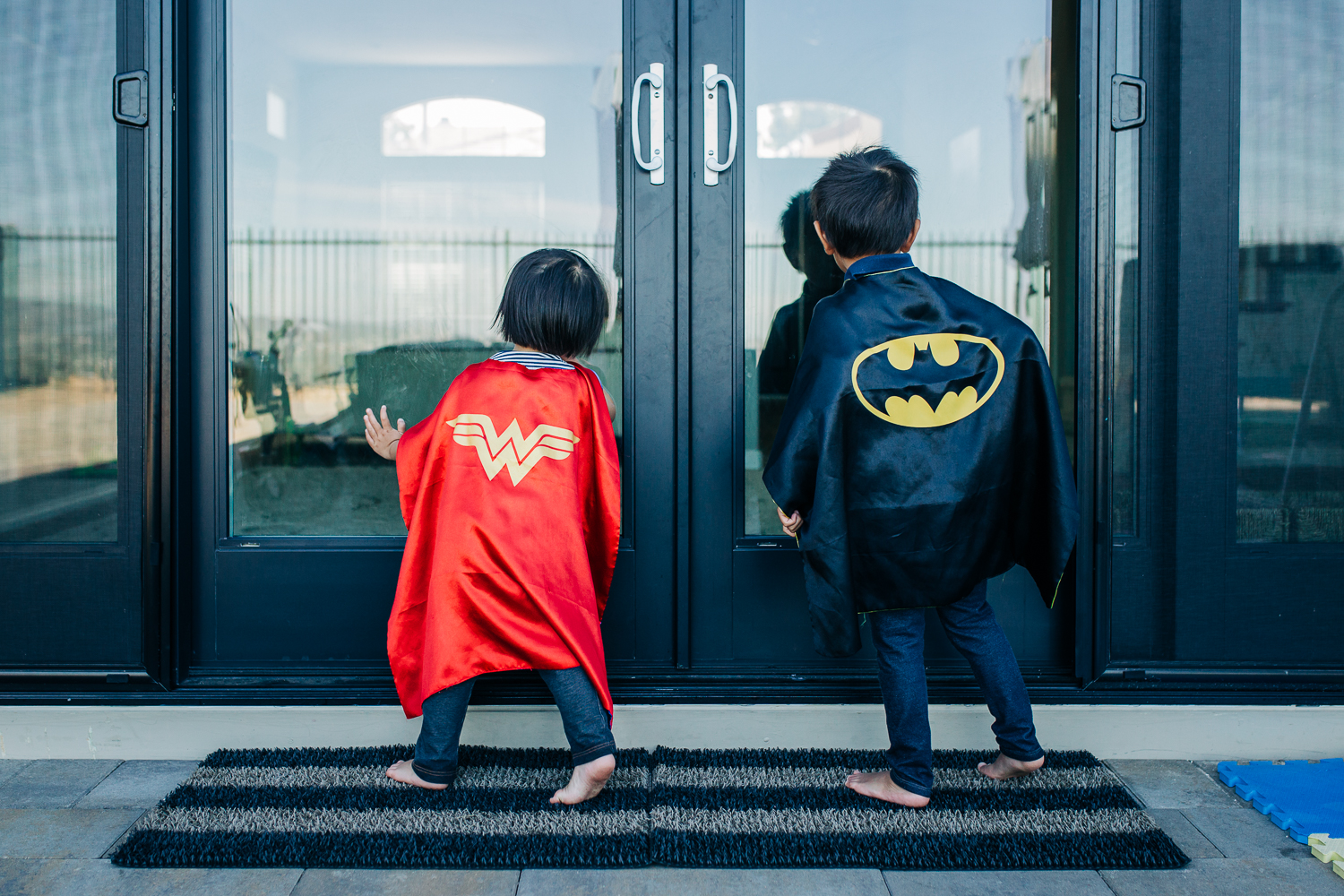 the backs of a boy and a girl wearing superhero capes as they look into sliding glass doors 