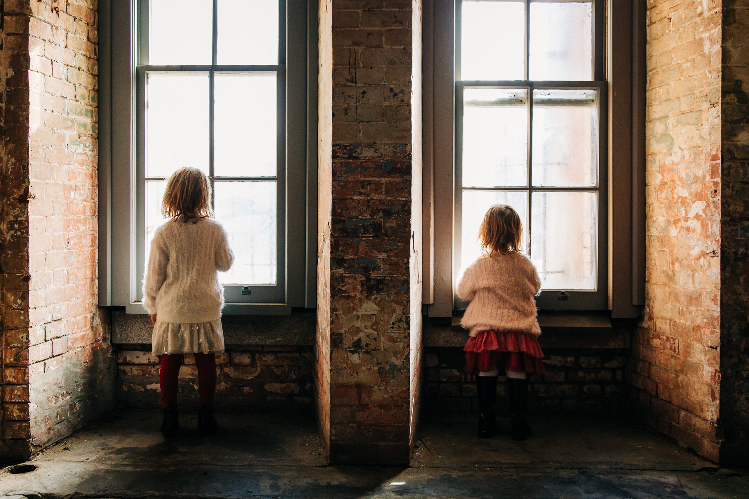 the back of two young sisters standing side next to each other looking out two separate windows at Fort Point