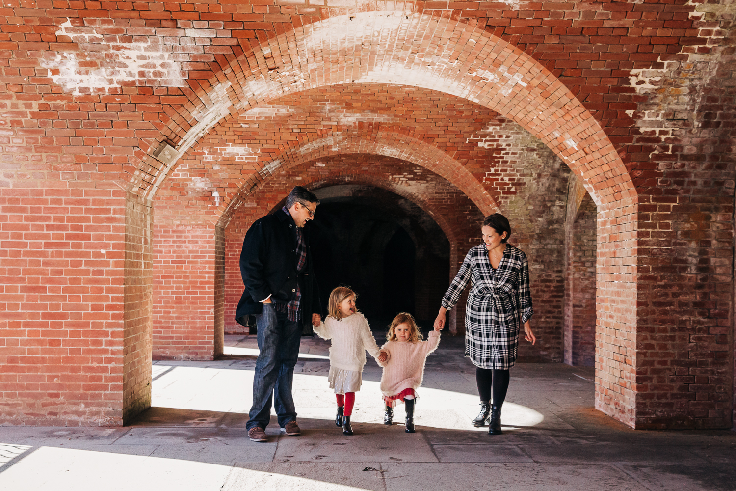 Family holding hands and walking together through a brick archway at Fort Point while looking at each other 