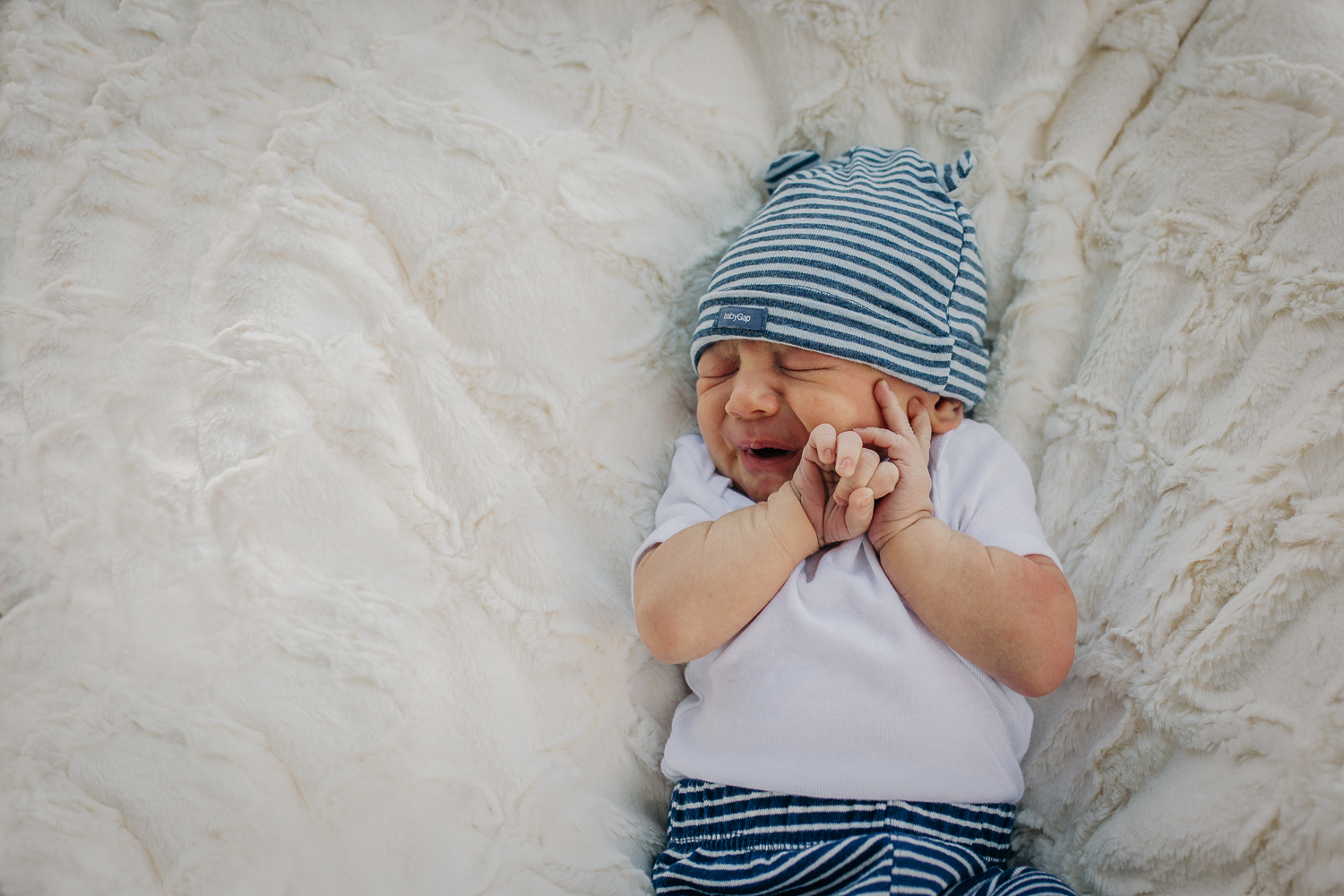 newborn boy laying down on a white blanket with his hands up by his face 