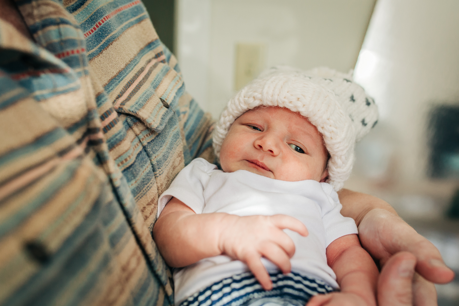 one week old baby boy in dad's arms looking towards camera while wearing a hat 