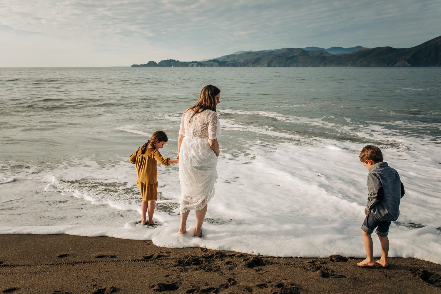 Pregnant mom playing at the beach in the water with her two children while wearing a white flowing dress 