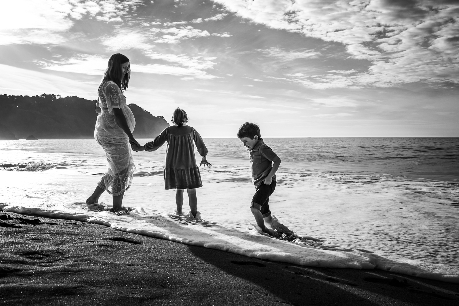 two small children playing in the water at the beach with their pregnant mother who is wearing a long white dress