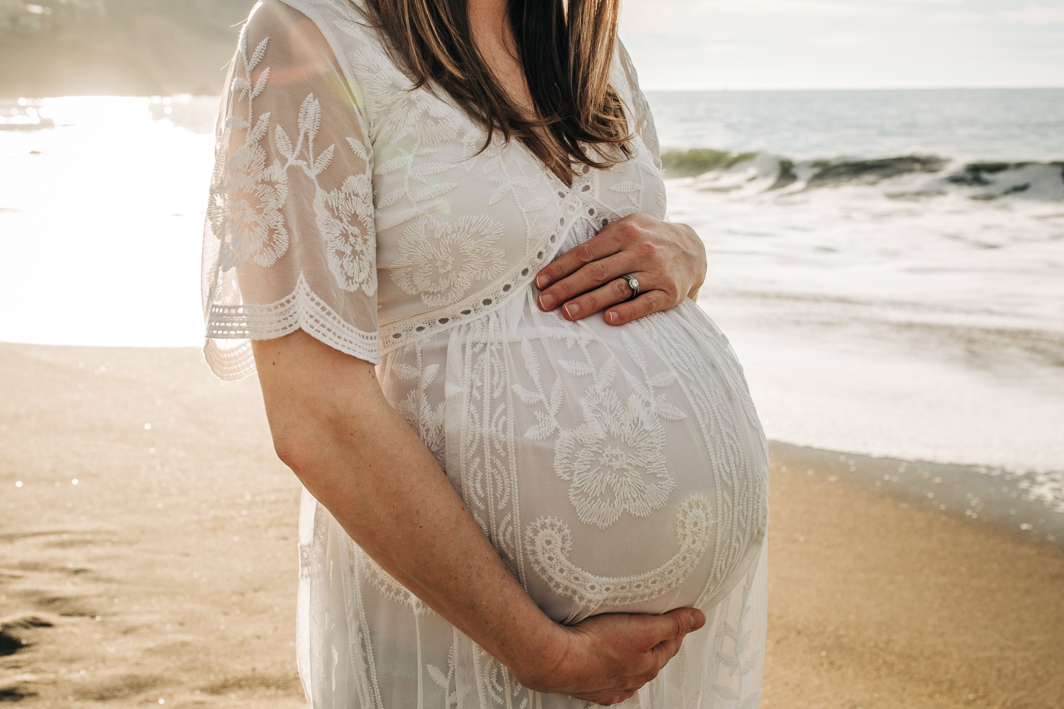 mother holding her baby bump while standing on the beach wearing a long white dress 