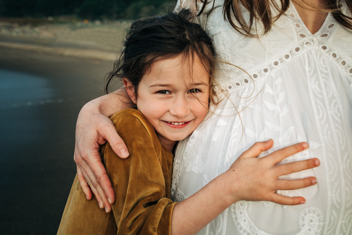 little girl smiling at the camera and snuggling her pregnant mother close and touching her mother's baby bump 