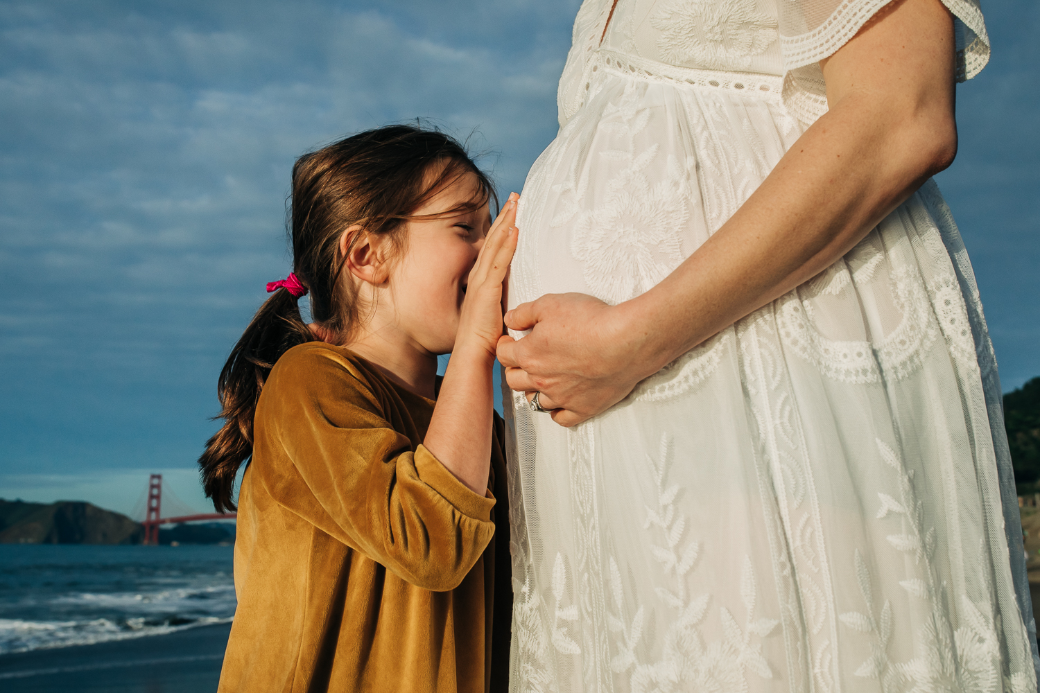 Little girl kissing her mother's baby bump at the beach with the golden gate bridge in the background 