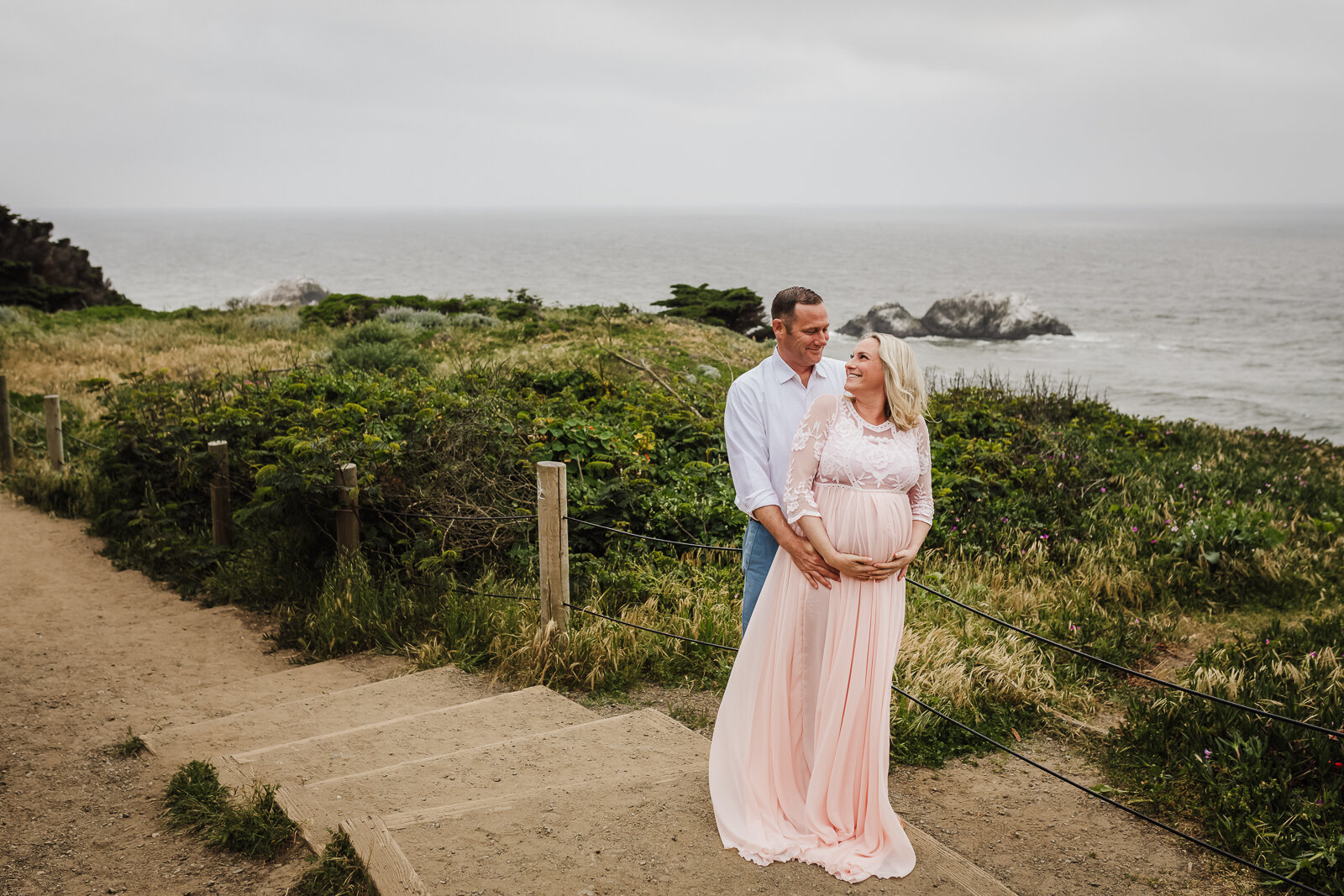 Husband standing behind pregnant wife and embracing her while standing along the coast in San Francisco