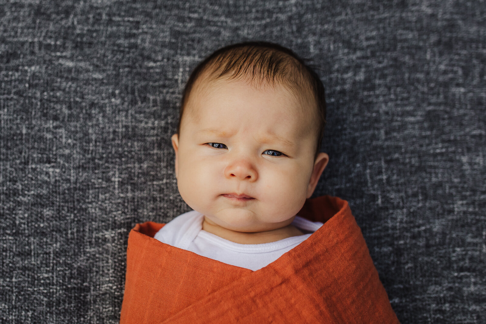 newborn swaddled in orange blanket and laying on gray blanket looking at camera 