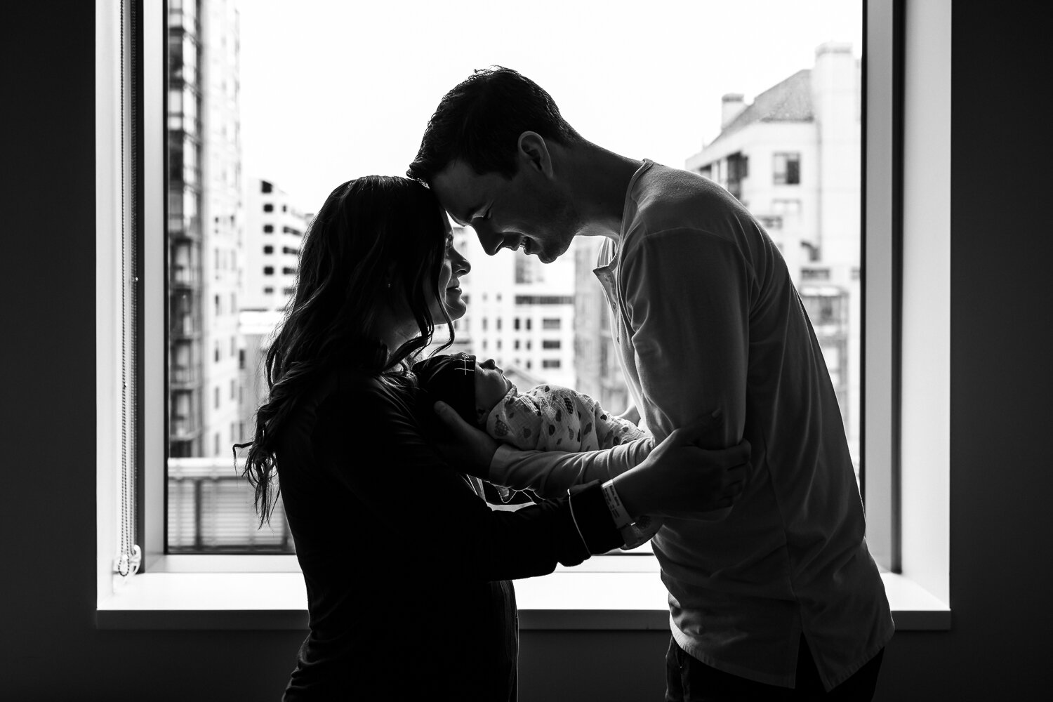 mom and dad standing in hospital window holding swaddled newborn together and touching foreheads smiling