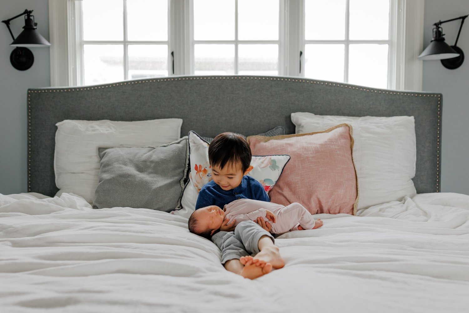 toddler boy holding newborn sister and looking down at her on parents' bed