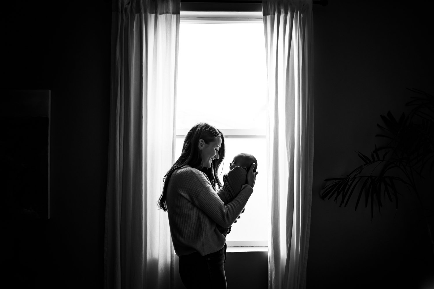 mom standing in front of window with curtains holding baby up to her face and smiling at her