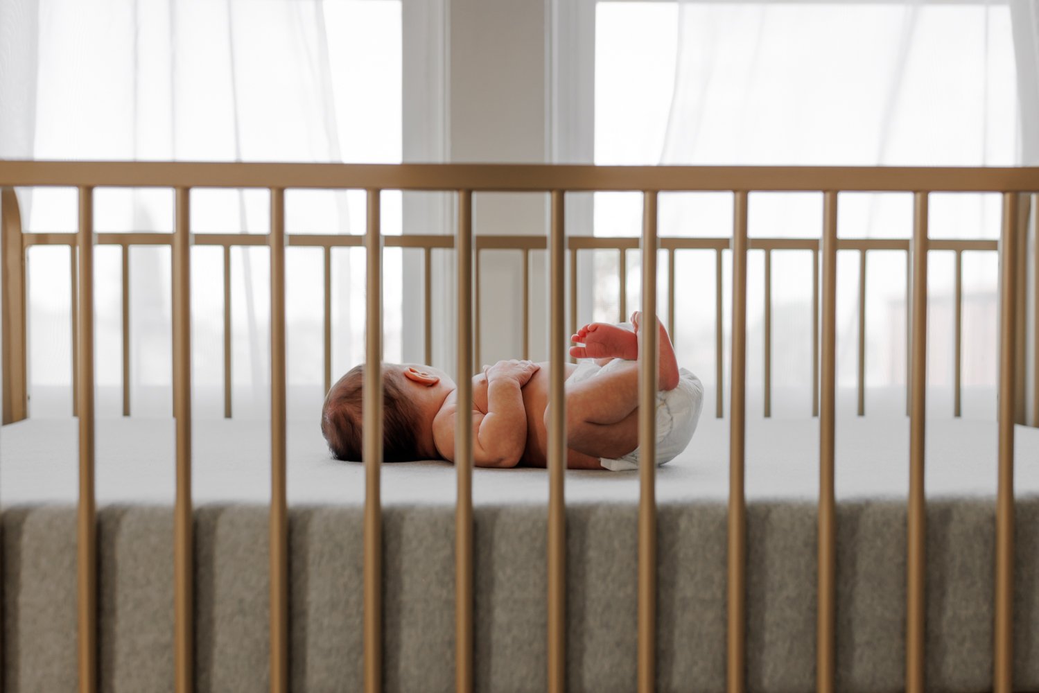 newborn in diaper laying in crib in front of windows with light curtains