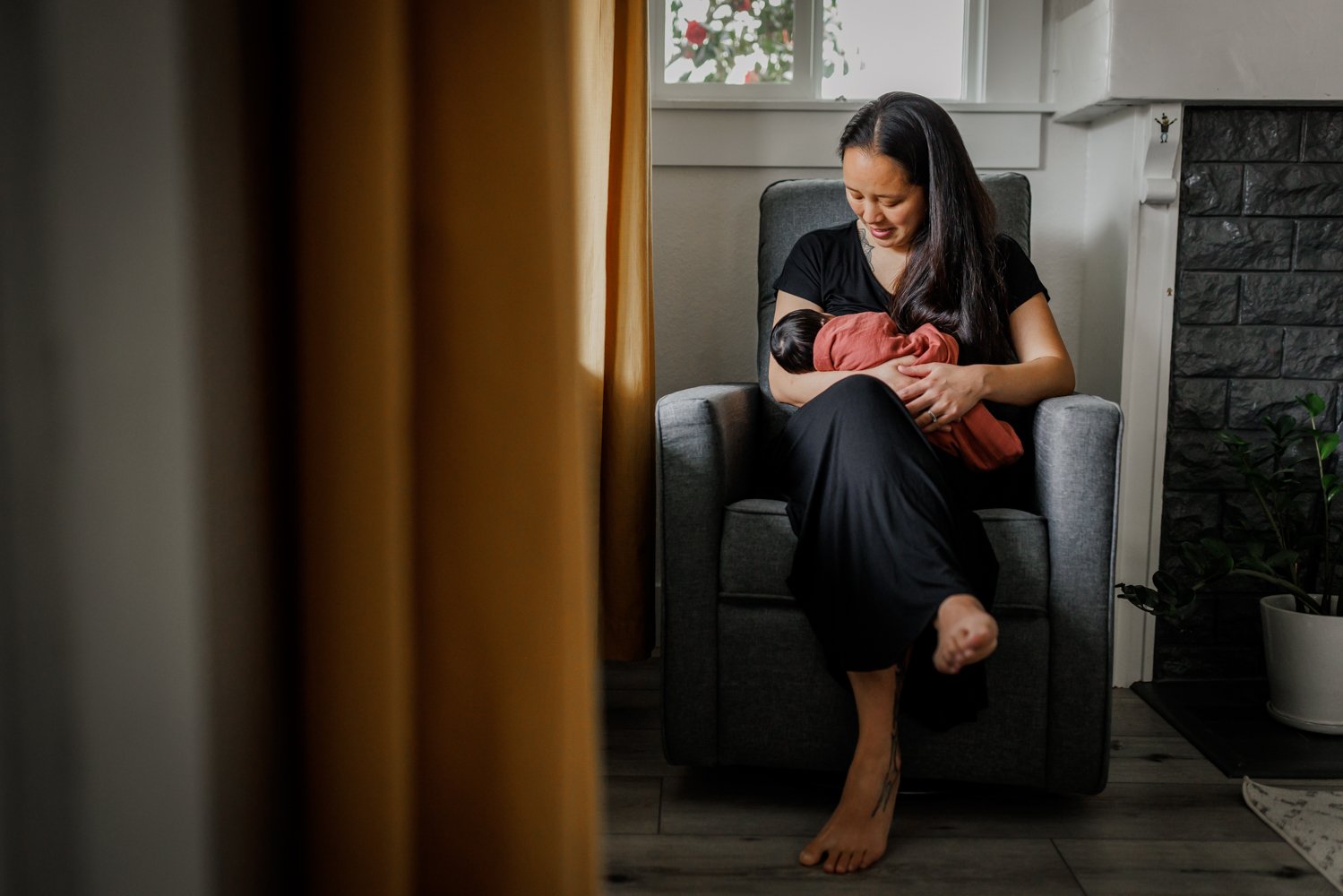 mother in black dress sitting by window nursing swaddled newborn