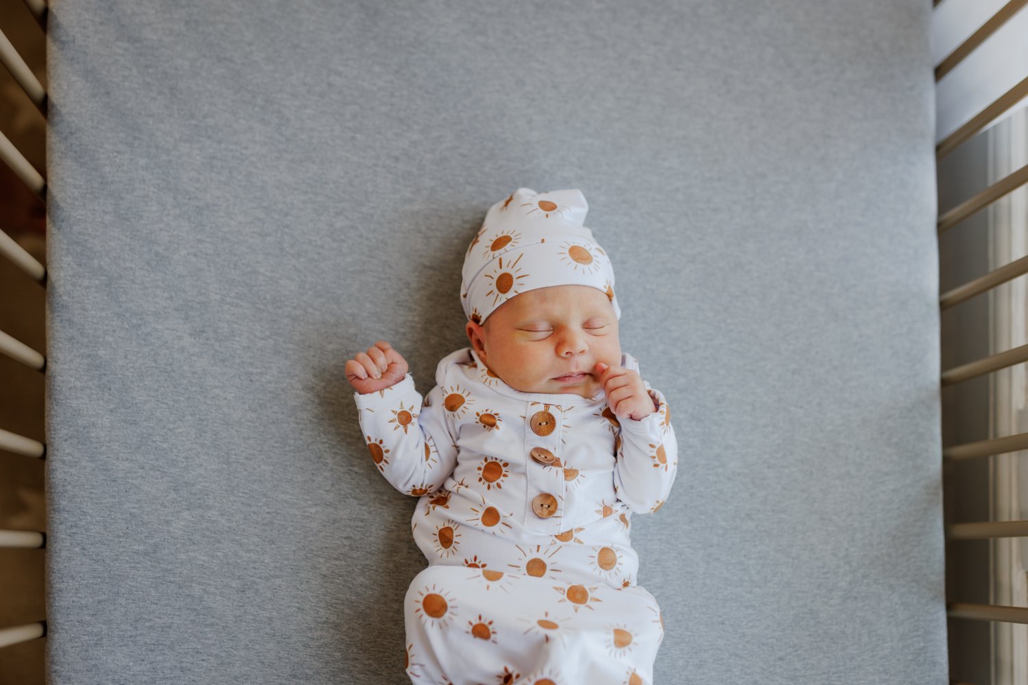 newborn in outfit with matching hat laying on gray sheet in crib