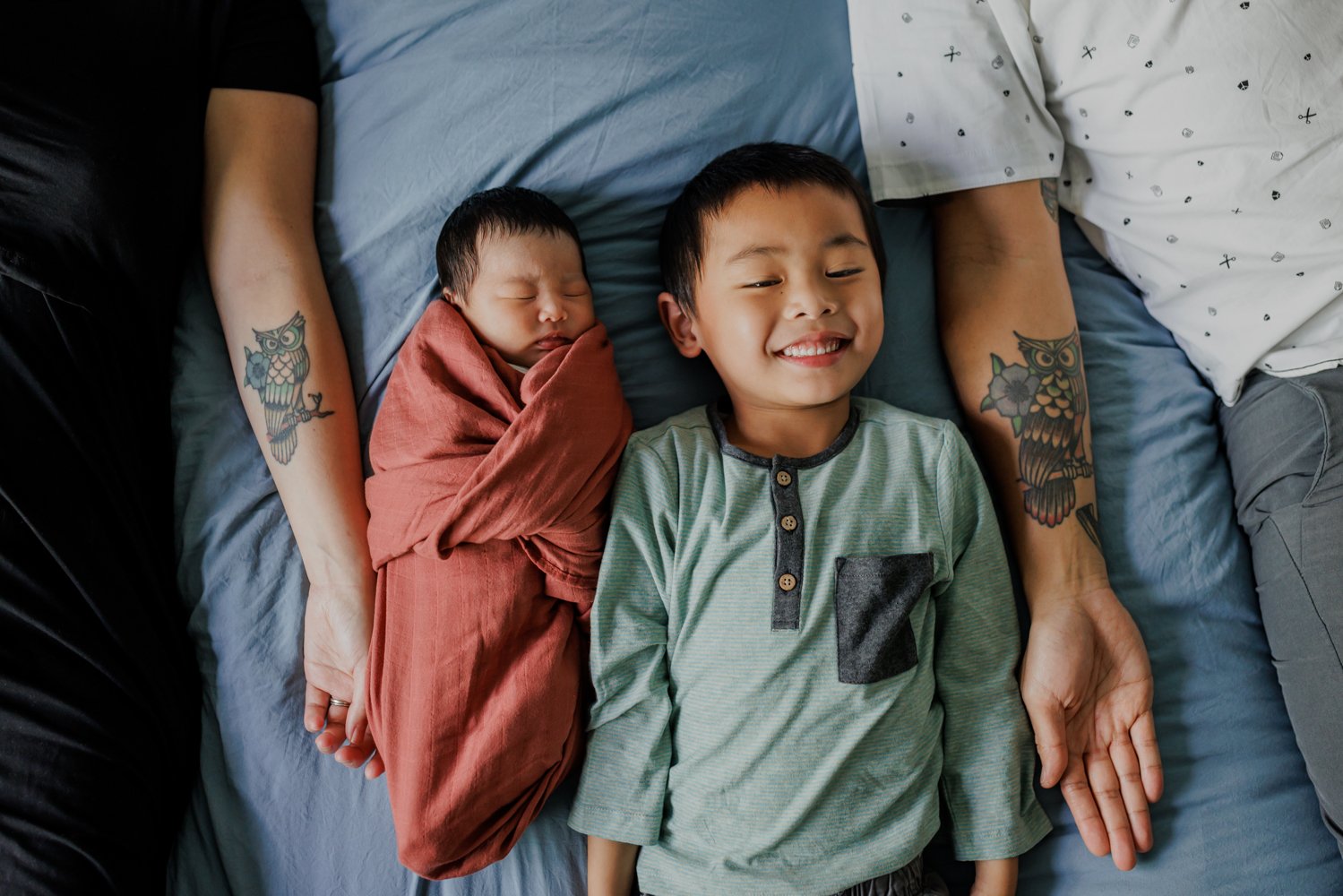 boy laying in bed next to swaddled newborn sister in between mom and dad's arms with matching owl tattoos