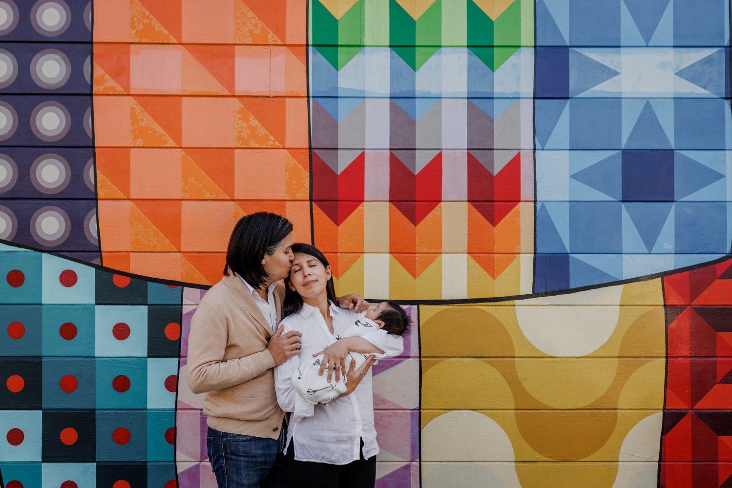 mom holding newborn in front of colorful San Francisco mural while husband hugs and kisses her