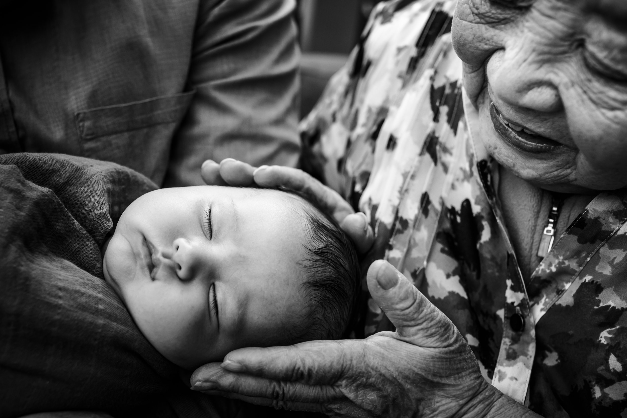 newborn with his hand held in his great grandmother's hands as she looks down at him and smiles at home