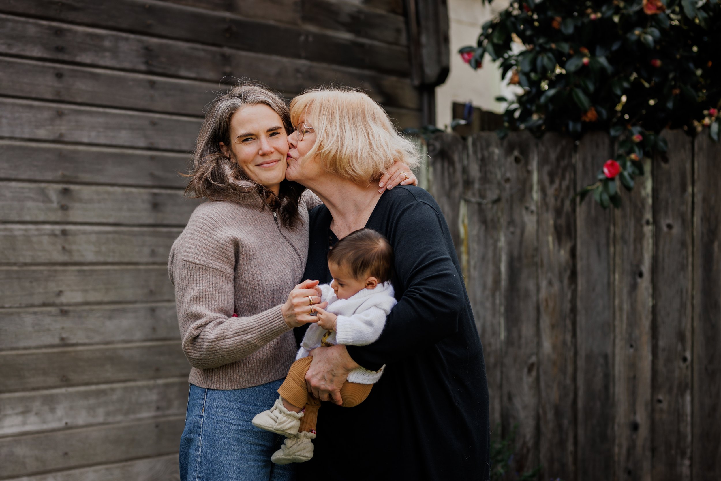 grandmother holding newborn grandaughter and leaning over to kiss her daughter who is smiling at the camera during Alameda session.jpg