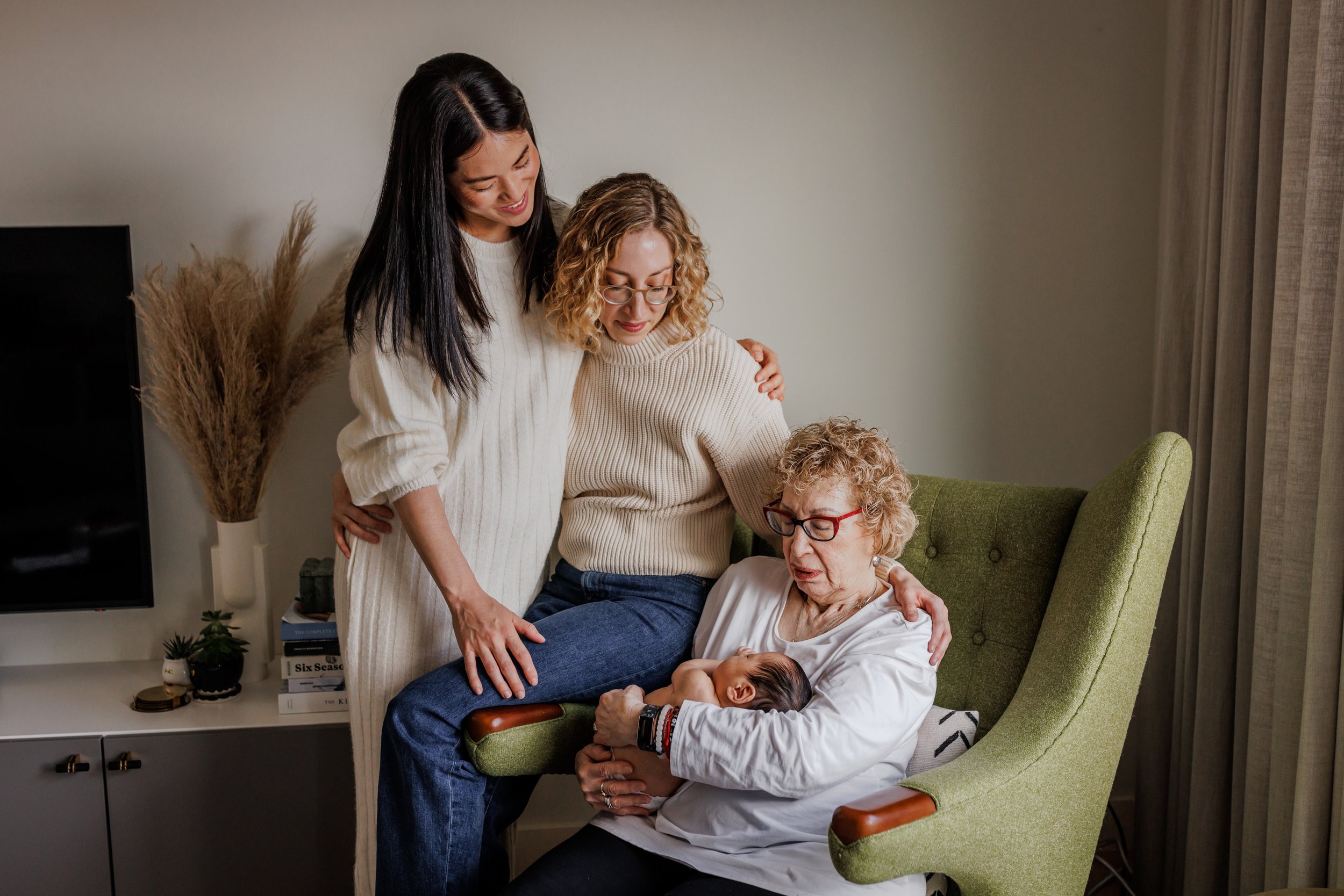 grandmother sitting in green chair holding baby grandson while his two moms look on during LGBTQ+ San Francisco in-home session.jpg