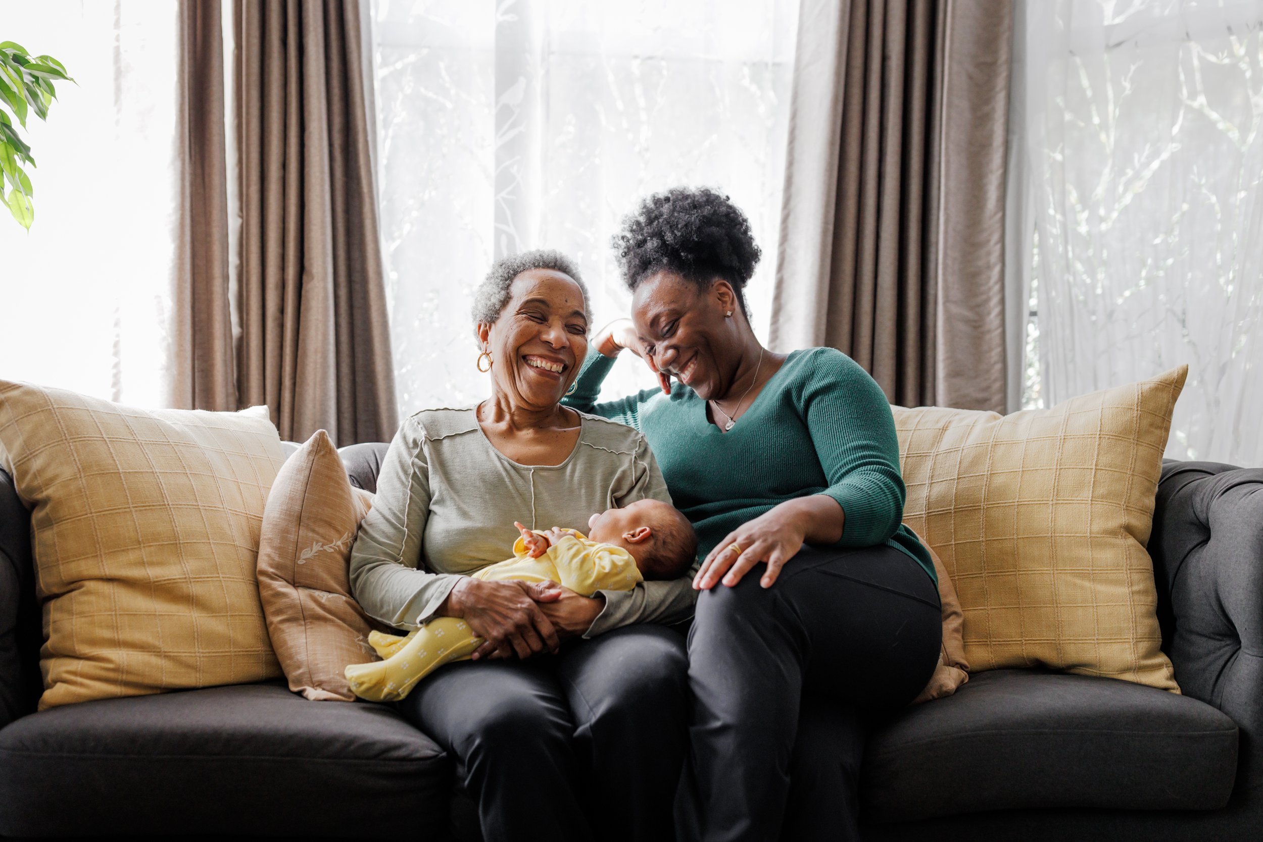 grandmother sitting on couch holding her newborn grandaughter next to her daugther while both smile during Oakland in-home session.jpg