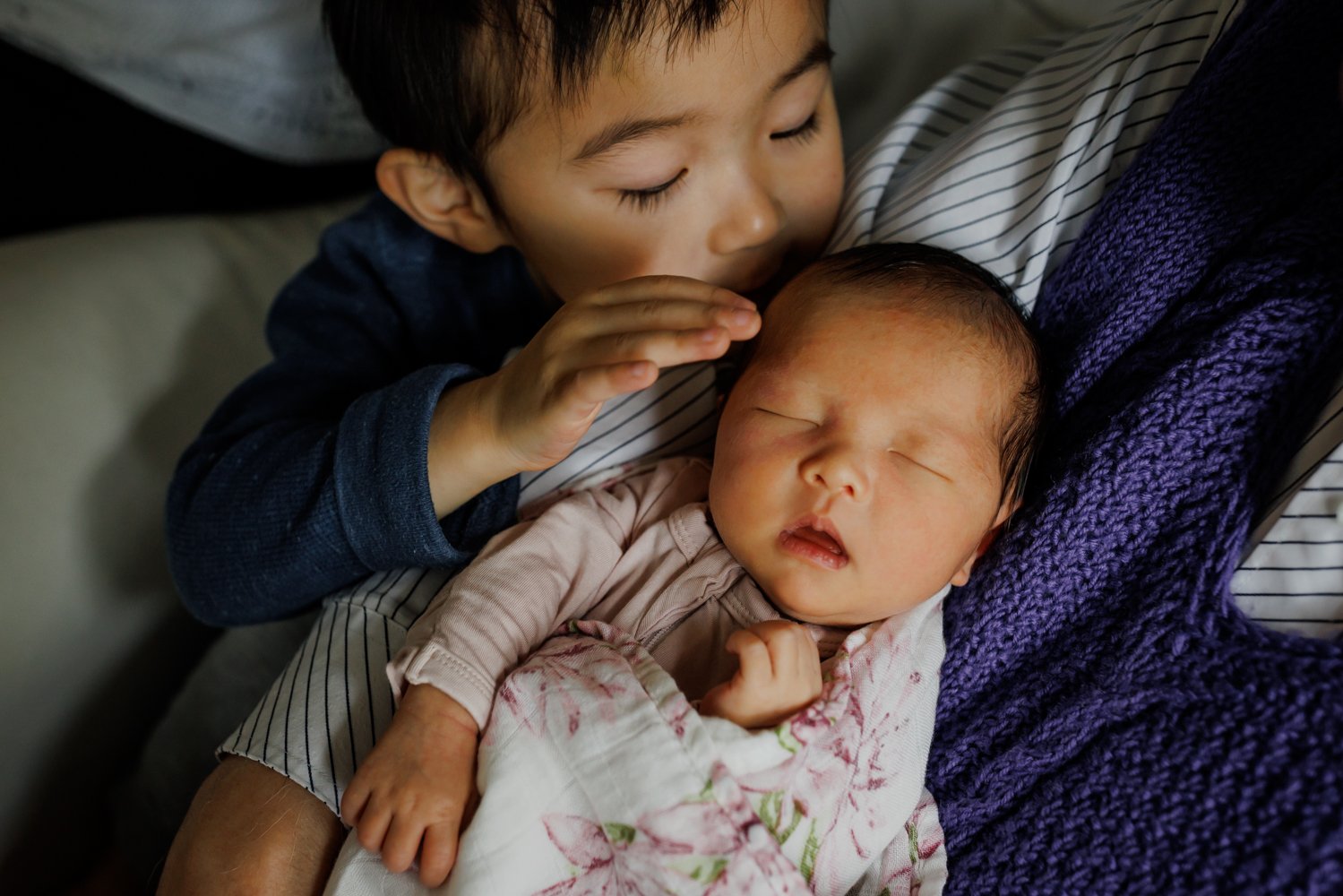 4-year-old boy gently touching and kissing his baby sibling's forehead while seated in grandpa's lap