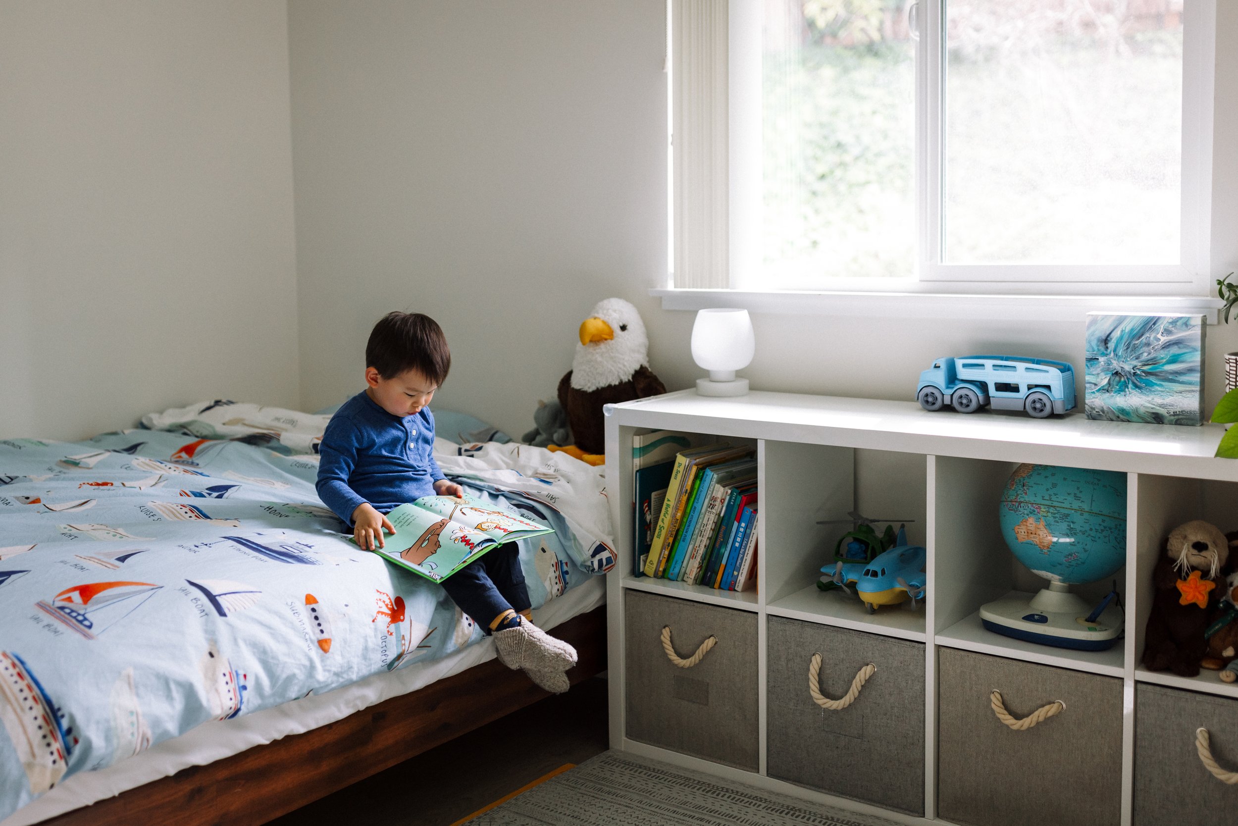 3-year-old boy sitting on the edge of his bed next to a white bookshelf reading