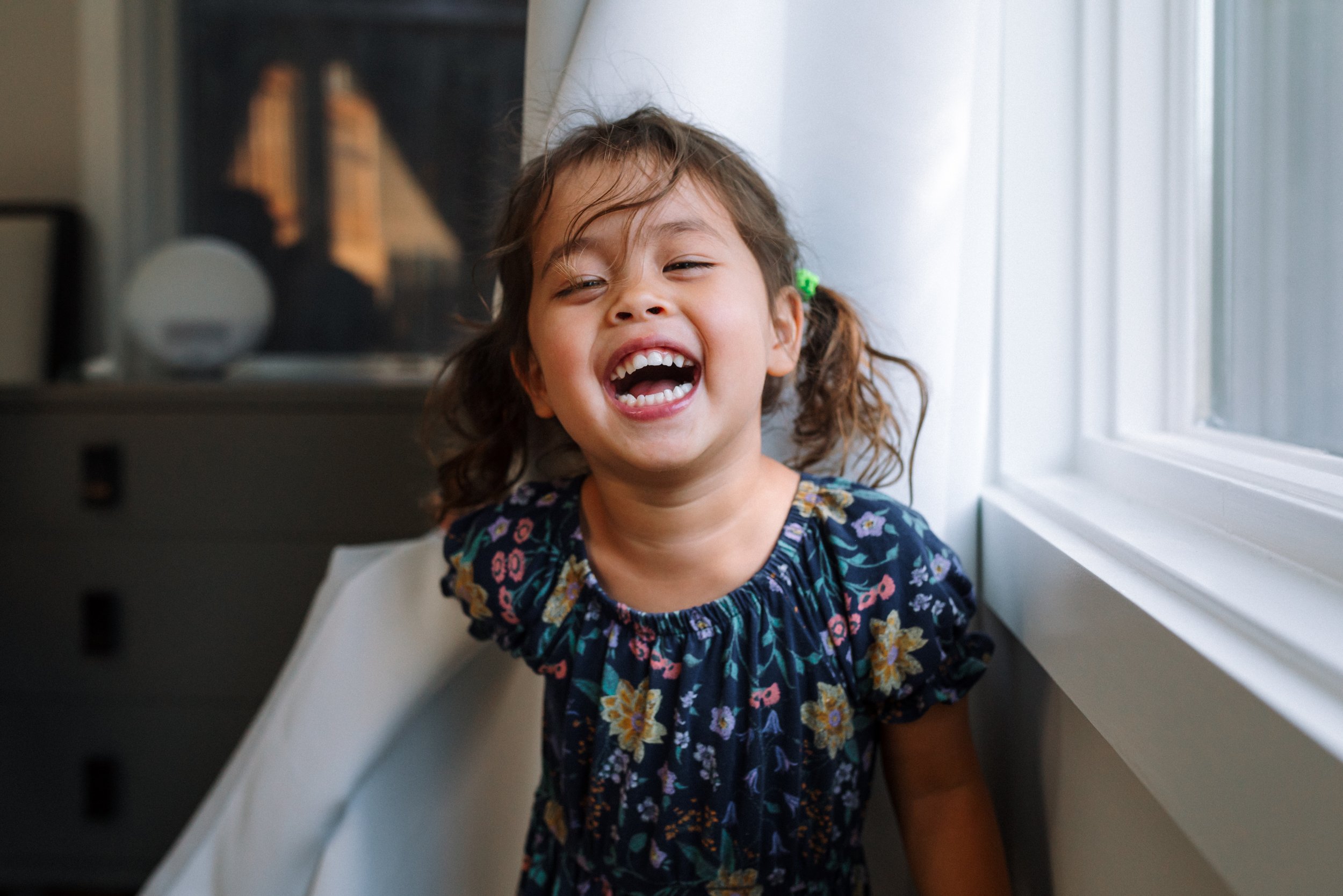 preschool-aged-girl-looking-at-camera-and-laughing-while-standing-next-to-window-at-home