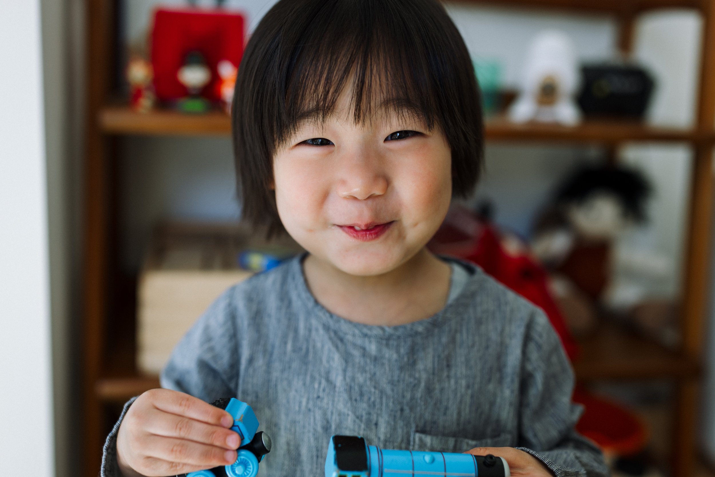 4-year-old boy smiling and looking at the camera in front of shelf while holding a blue thomas train