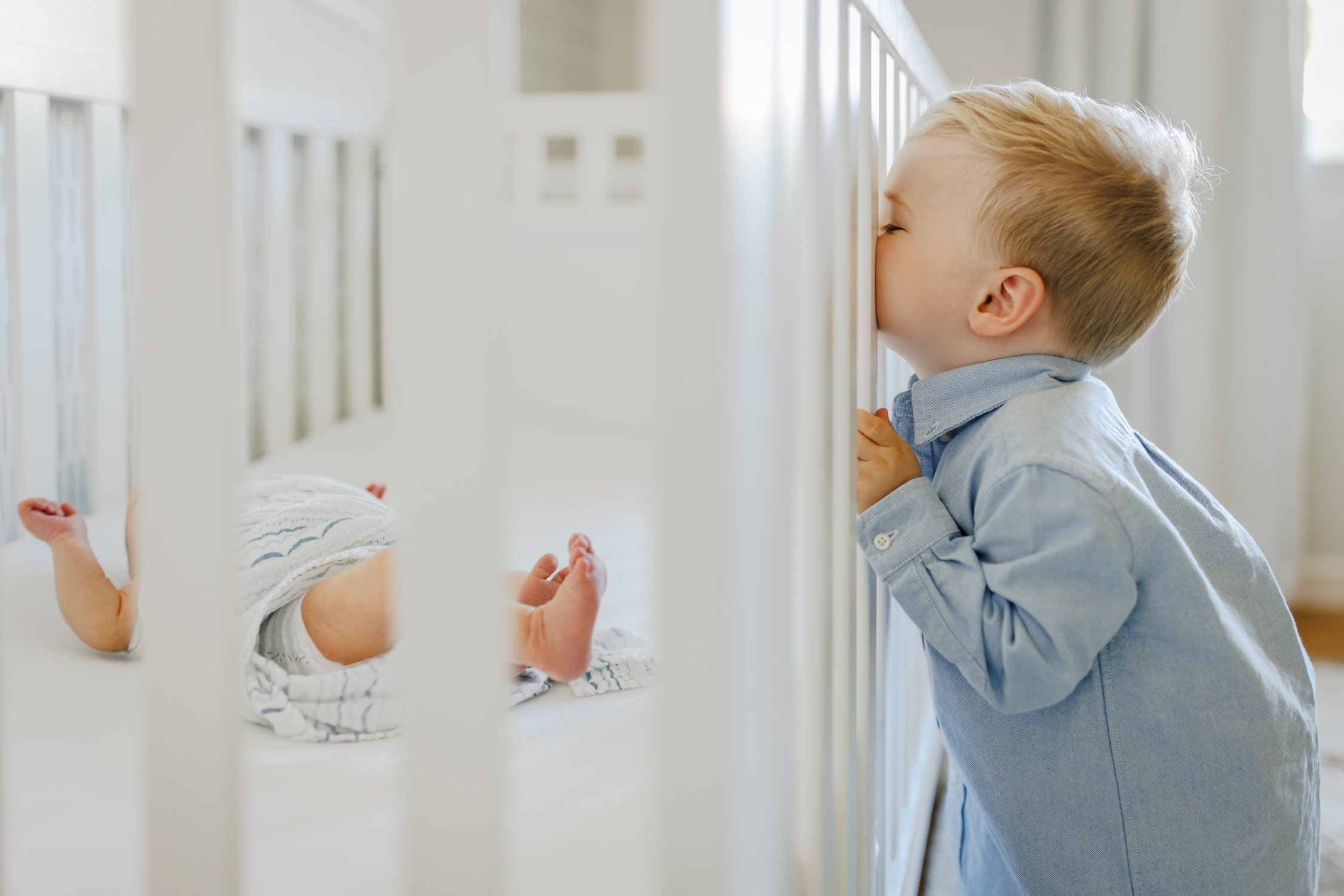 toddler boy peeking into white crib to see his newborn brother