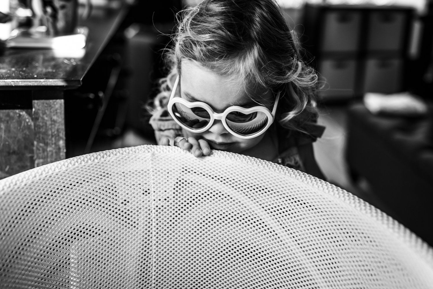 black and white photo of a toddler girl wearing heart sunglasses peering into her newborn sister's bassinet