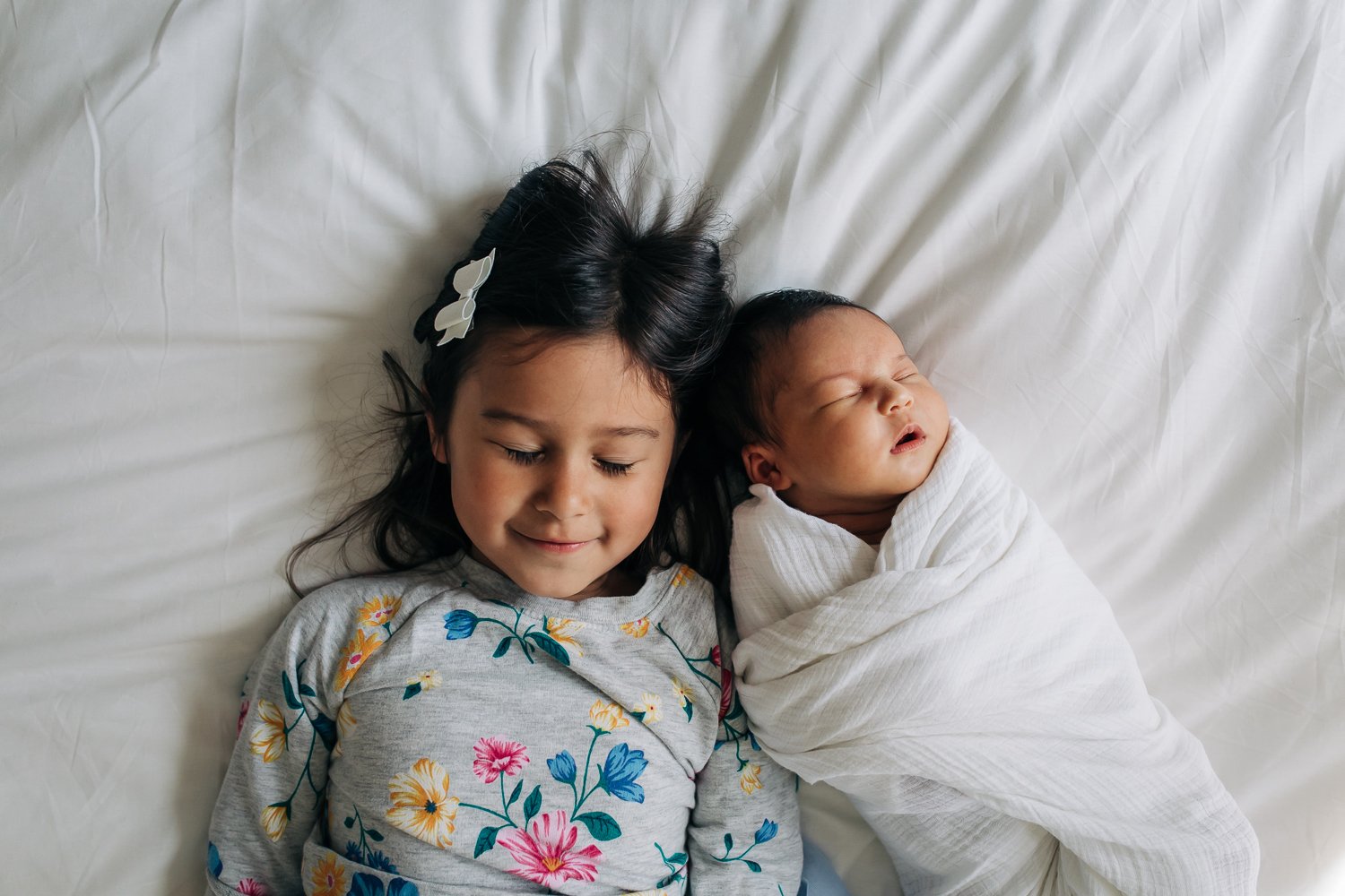 sister and newborn sibling laying together on a bed with a white comforter with their eyes closed