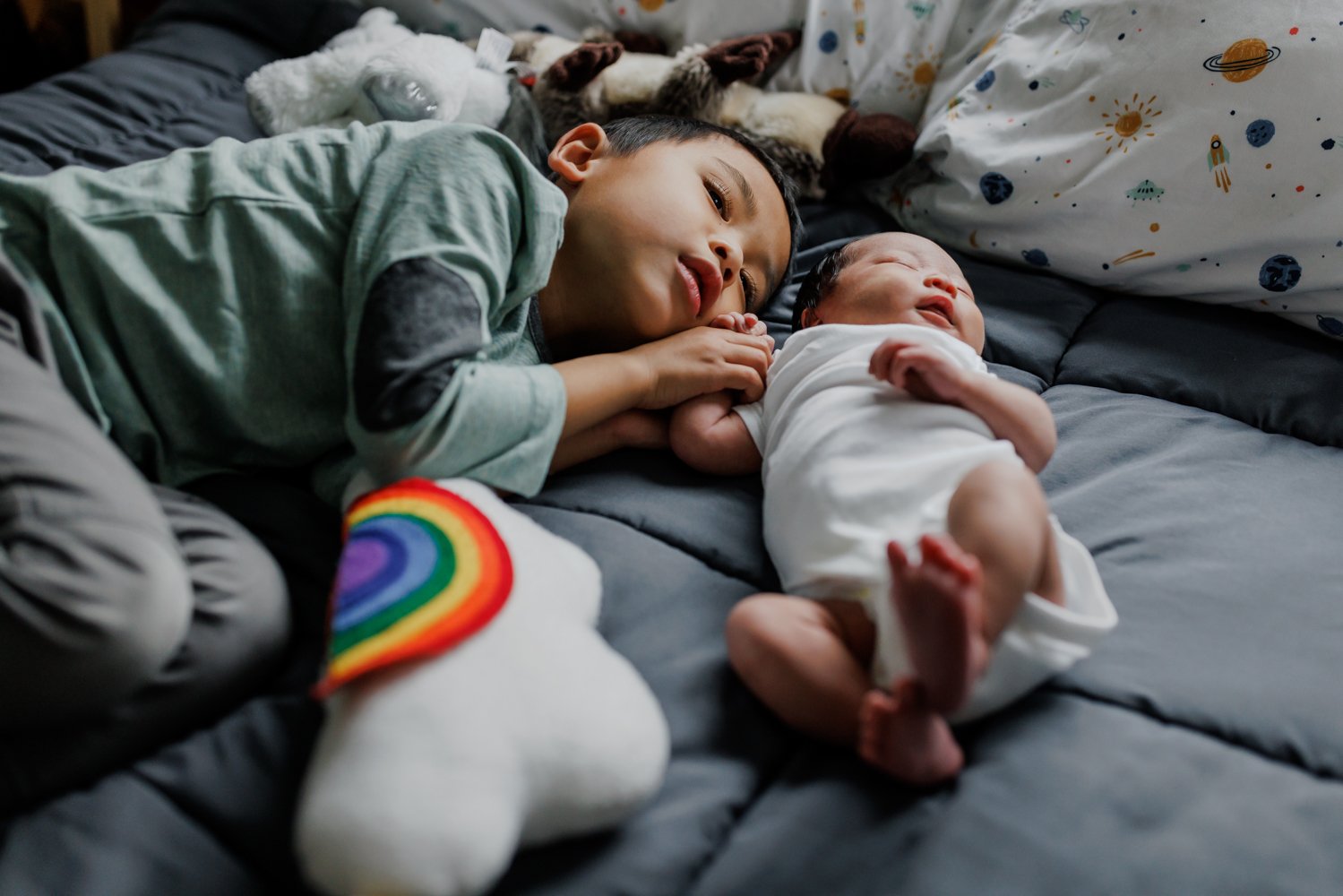 preschool-aged boy laying on a bed next to newborn brother with a rainbow pillow in the foreground