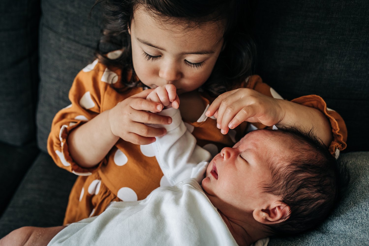 toddler sister holding her newborn brother in her lap and gently kissing his hand