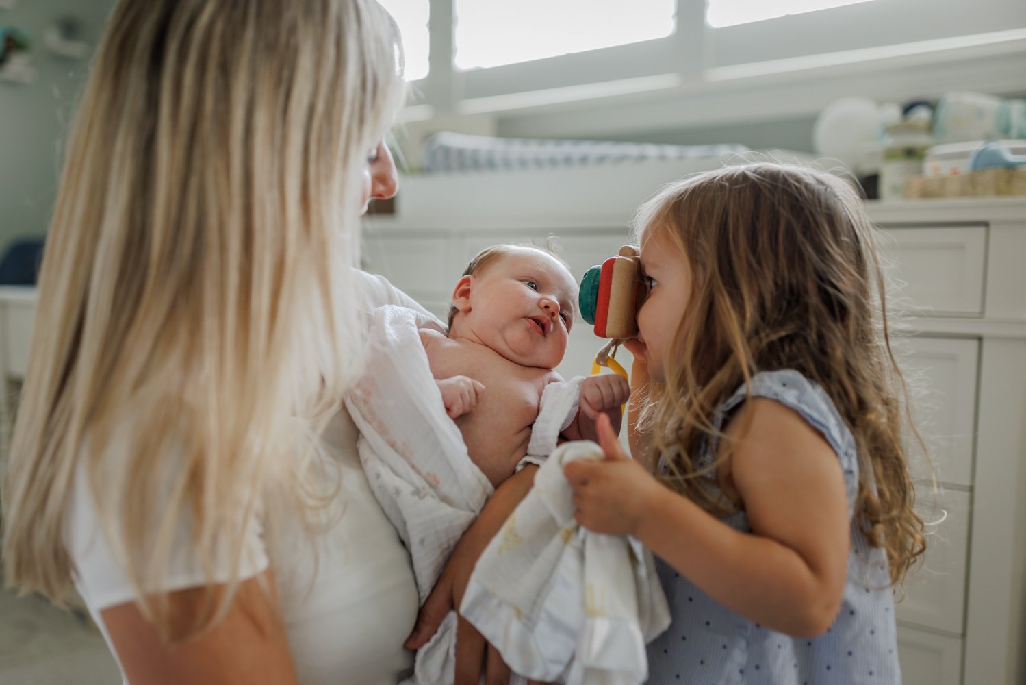 toddler girl pretending to take a photo of her baby sister with her toy camera while the mom holds the baby in her arms