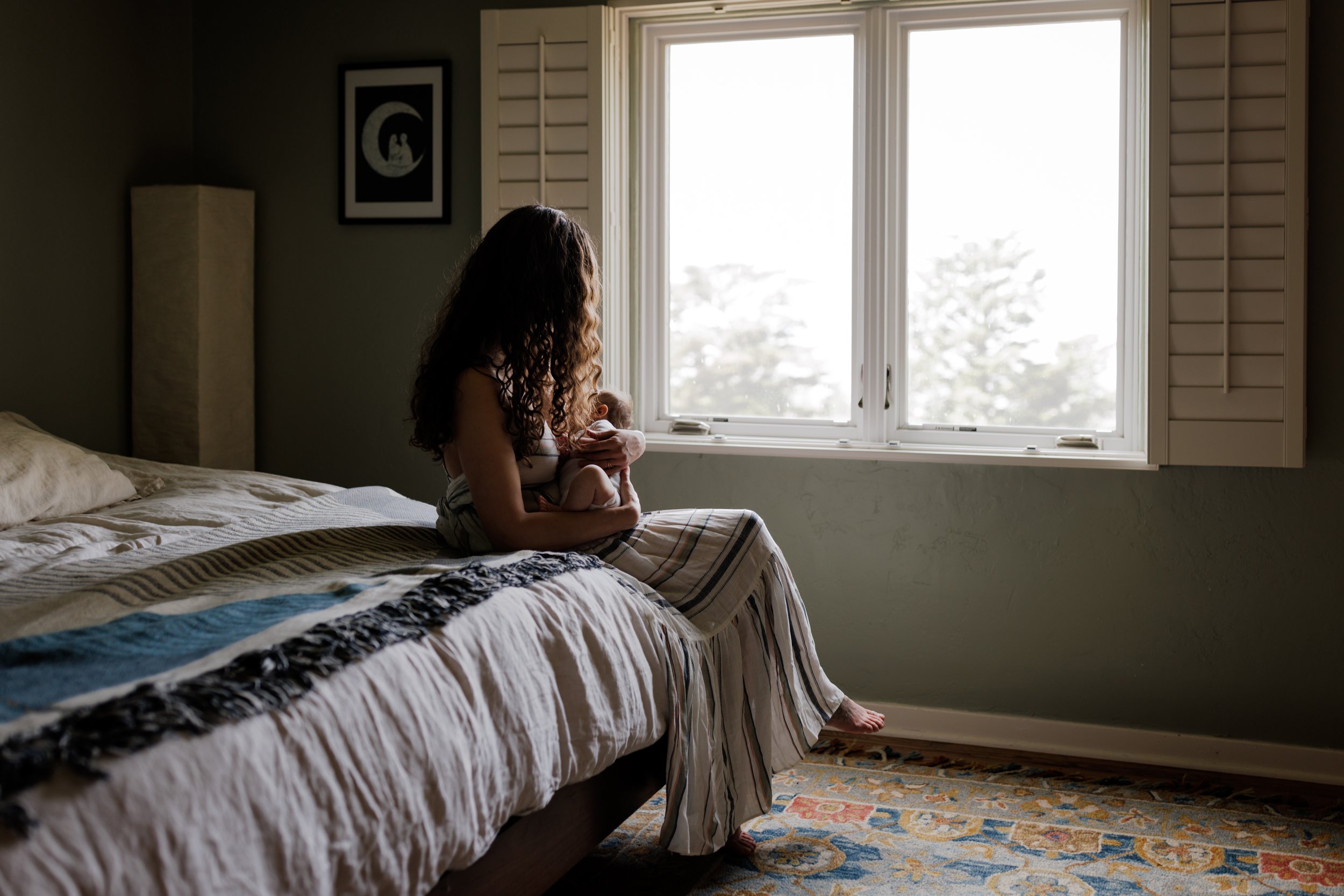 Mom sitting on edge of bed my large window looking away and holding newborn