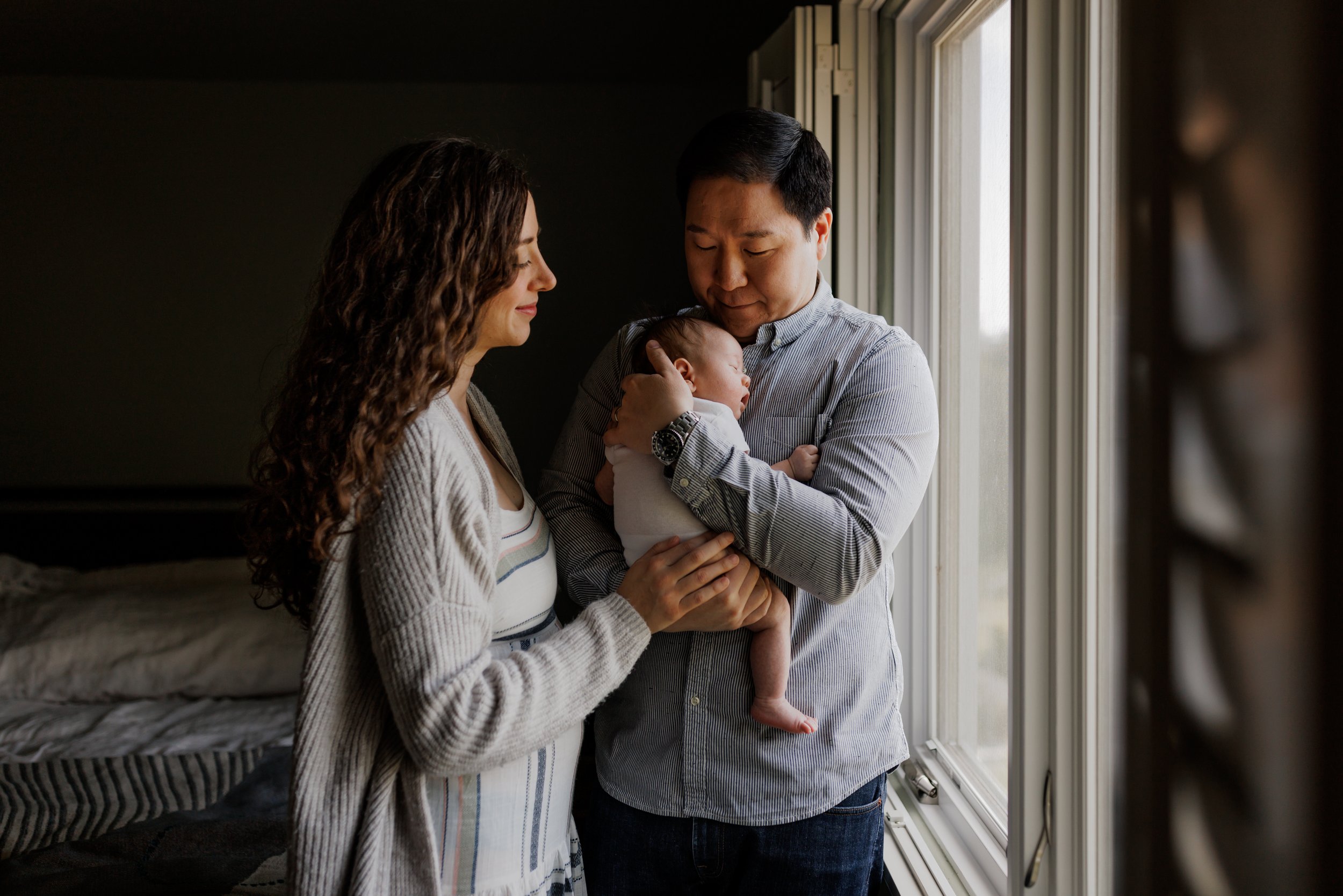 parents standing next to window in bedroom with dad holding baby and mom resting hand on dad's hand